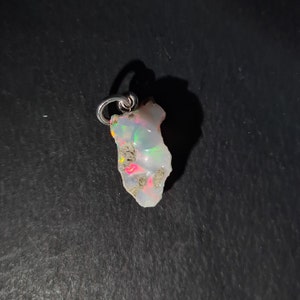 Op de afbeelding: Een witte opaal hanger met een zilveren beugel. De opaal heeft een ruwe, natuurlijke vorm en vertoont iriserende kleuren.