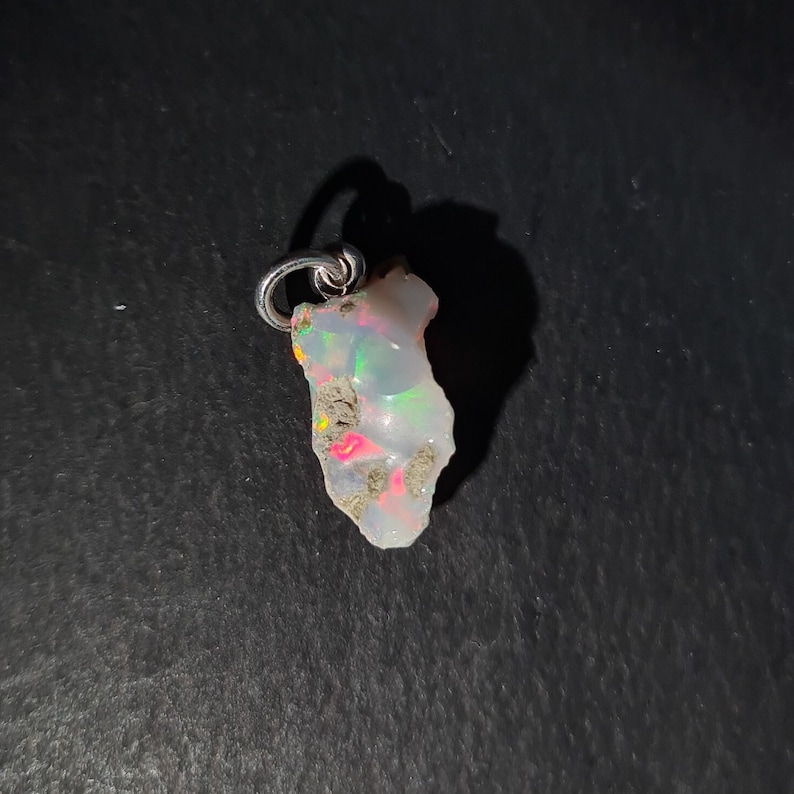 Op de afbeelding: Een witte opaal hanger met een zilveren beugel. De opaal heeft een ruwe, natuurlijke vorm en vertoont iriserende kleuren.