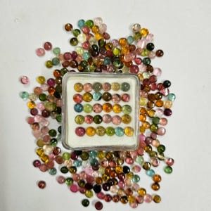 Op de afbeelding: Een verzameling kleurrijke, ronde edelstenen in verschillende tinten roze, groen, geel en bruin. Sommige edelstenen zijn gerangschikt in een doorzichtige plastic doos, andere zijn verspreid op een wit oppervlak. De edelstenen zijn gepolijst.