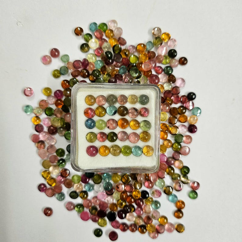 Op de afbeelding: Een verzameling kleurrijke, ronde edelstenen in verschillende tinten roze, groen, geel en bruin. Sommige edelstenen zijn gerangschikt in een doorzichtige plastic doos, andere zijn verspreid op een wit oppervlak. De edelstenen zijn gepolijst.
