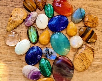 May include: Assortment of polished gemstone cabochons in various colors and shapes, including green, blue, red, brown, and purple. The stones are arranged in a heart shape on a wooden surface.