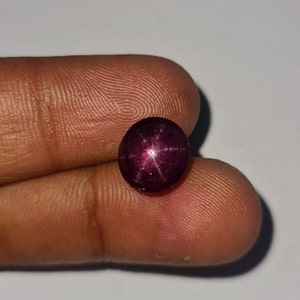 Op de afbeelding: Een ronde, gepolijste, donkerrode edelsteen met een zesstralig sterpatroon. De edelsteen wordt tussen vingers gehouden, waarbij de diepe kleur en het stereffect, gecreëerd door lichtreflectie in de steen, worden getoond. De edelsteen is een cabochon-slijpsel.