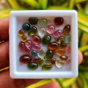 May include: A white box filled with a variety of small, colorful gemstones. The gemstones are in various shapes and sizes, including round, oval, and teardrop. The gemstones are a mix of pink, green, blue, and brown.