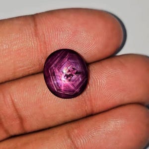 Puede incluir: Una gema ovalada de color púrpura oscuro con efecto estrella. La piedra pulida muestra un patrón de estrella de seis puntas que irradia desde el centro. La gema se sostiene en una mano, resaltando su color y claridad.