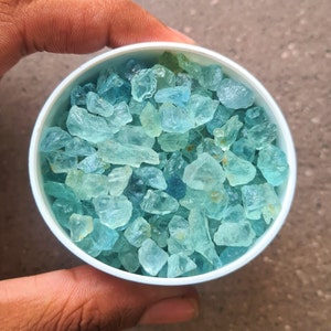 May include: A white bowl filled with light blue aquamarine gemstones. The bowl is being held by a hand.