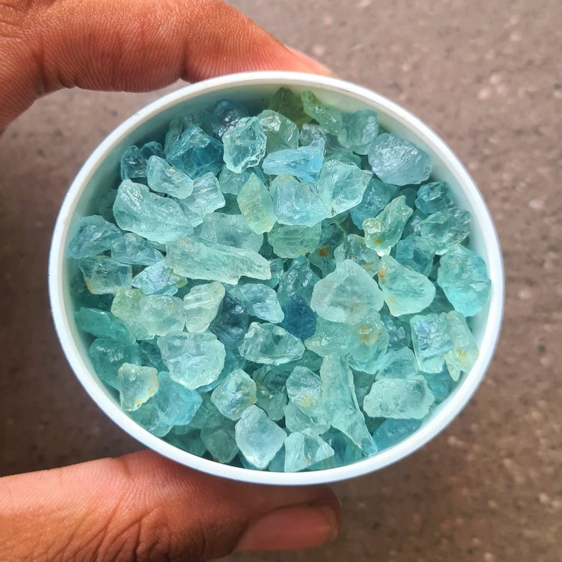 May include: A white bowl filled with light blue aquamarine gemstones. The bowl is being held by a hand.