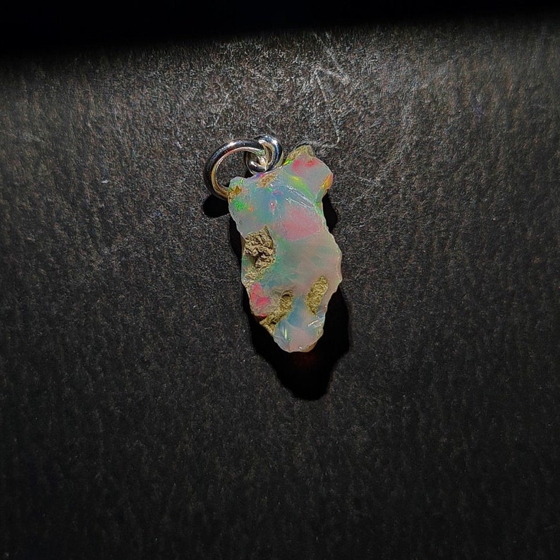 Op de afbeelding: Een zilveren hanger met een ruwe, iriserende opaal met een onregelmatige vorm. De opaal heeft een mix van witte, roze en blauwe kleuren.