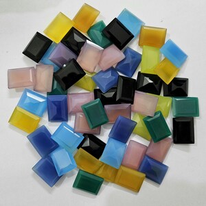 Könnte beinhalten: Sortiment aus bunten rechteckigen Edelstein-Cabochons in verschiedenen Farbtönen von Blau, Grün, Gelb, Rosa, Lila und Schwarz. Die Steine sind in einem zufälligen Muster auf einer weißen Oberfläche angeordnet.