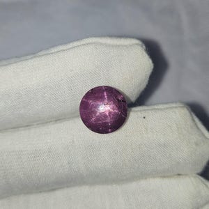 May include: A round, purple gemstone with a star-like pattern in the centre. The gemstone is set against a white background.