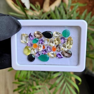 Puede incluir: Una colección de piedras preciosas sueltas en varias formas, incluyendo cortes ovalados, de pera, redondos y rectangulares. Las piedras presentan una paleta de colores diversa de violeta, amarillo, verde, naranja, negro y tonos claros, exhibidas en una bandeja blanca.