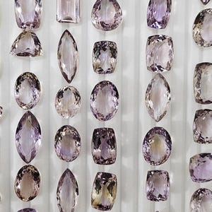 Könnte beinhalten: Eine Anordnung von Amethyst-Edelsteinen in verschiedenen Formen und Farbtönen von Lila. Die Edelsteine sind vor einem weißen Hintergrund angeordnet und zeigen verschiedene Formen wie oval, rechteckig und tropfenförmig. Die Steine sind in einem Raster angeordnet.