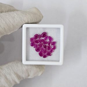 May include: A collection of polished, vibrant pink-red gemstones in various shapes, including oval and teardrop, displayed in a white square container. The gemstones are arranged on a white surface, creating a visually appealing display.
