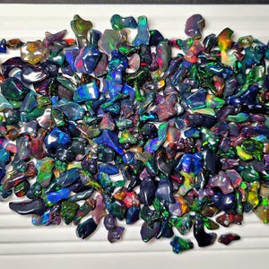 May include: A collection of raw, uncut opals in various shapes and sizes. The opals display a vibrant play of color, with flashes of blue, green, red, and purple. The gemstones are scattered across a white surface, showcasing their natural beauty.