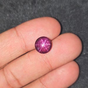 Könnte beinhalten: Ein runder, tiefroter Edelstein mit einem sternartigen Muster auf seiner Oberfläche. Der Edelstein wird in einer Hand gehalten, wodurch seine polierten, reflektierenden Facetten hervorgehoben werden. Der Sterneffekt wird durch Einschlüsse im Stein erzeugt.