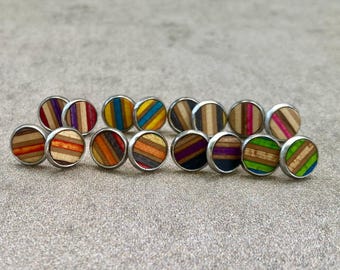 Könnte beinhalten: Eine Sammlung runder Ohrstecker mit geschichteten, mehrfarbigen Holzmusterungen. Jedes Paar zeigt markante Streifendesigns in verschiedenen Farben wie Rot, Blau, Lila, Grün, Orange und Naturholztönen, eingefasst in silberfarbenes Metall.