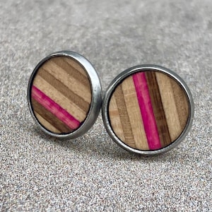 K&ouml;nnte beinhalten: Zwei runde Ohrstecker mit silberfarbenem Metallrand. Die Ohrringmitten weisen ein gestreiftes Design aus hellbraunem, dunkelbraunem und rosa Holz auf. Die Ohrringe sind vor einem strukturierten grauen Hintergrund platziert.