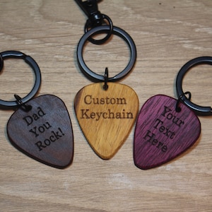 Puede incluir: Tres llaveros con forma de p&uacute;a de guitarra. Uno es marr&oacute;n oscuro con "Dad You Rock!" grabado, otro marr&oacute;n claro con "Custom Keychain", y el tercero morado con "Your Text Here". Cada uno tiene un anillo y un cierre de metal negro.