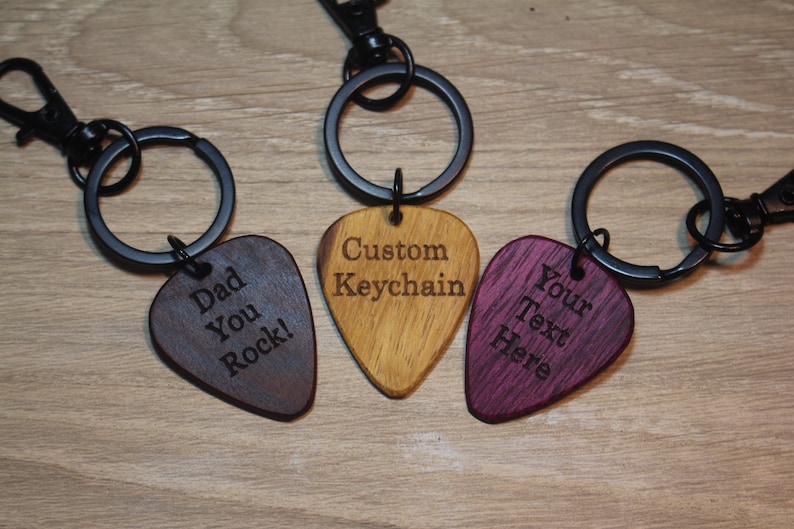 Puede incluir: Tres llaveros con forma de p&uacute;a de guitarra. Uno es marr&oacute;n oscuro con "Dad You Rock!" grabado, otro marr&oacute;n claro con "Custom Keychain", y el tercero morado con "Your Text Here". Cada uno tiene un anillo y un cierre de metal negro.