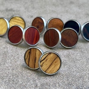 May include: A collection of circular stud earrings featuring various wood grain inlays set within silver-toned metal frames. The wood colors range from light yellow and tan to deep reddish-brown and dark black, showcasing natural textures and patterns.