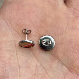 K&ouml;nnte beinhalten: Ein Paar Ohrstecker aus Edelstahl. Jeder Ohrring hat ein rundes Holzdesign mit rosa und braunen Streifen. Die Ohrringe haben einen Stecker und einen Schmetterlingsverschluss f&uuml;r sicheren Halt.