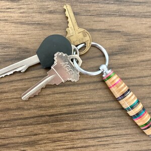 May include: A silver key chain with three keys and a wooden keychain charm. The keychain charm is made of wood with colorful stripes and a silver cap on the end. The keys are silver and have a black plastic head. The keychain is on a wooden surface.