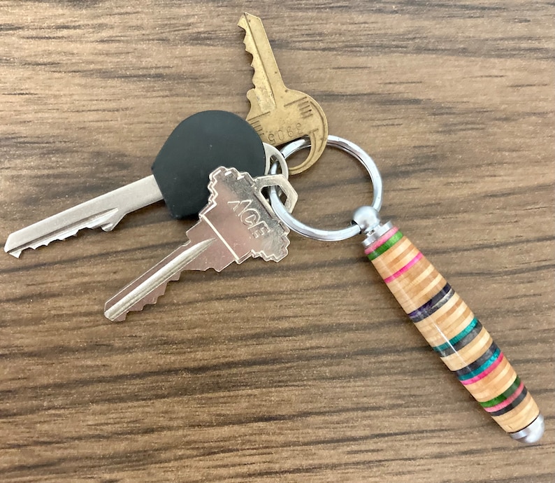 May include: A silver key chain with three keys and a wooden keychain charm. The keychain charm is made of wood with colorful stripes and a silver cap on the end. The keys are silver and have a black plastic head. The keychain is on a wooden surface.