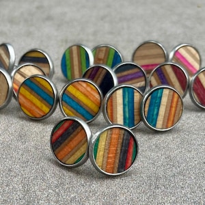 Puede incluir: Una colección de pendientes de botón redondos con un borde de metal plateado. Los pendientes presentan un diseño a rayas colorido hecho de madera, con varios colores como rojo, azul, amarillo y verde.
