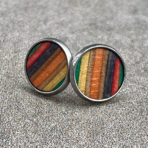 K&ouml;nnte beinhalten: Ein Paar runde Ohrstecker mit silberfarbenem Rand. Die Ohrringe weisen ein farbenfrohes Streifenmuster mit Rot-, Orange-, Gelb-, Gr&uuml;n- und Schwarzt&ouml;nen auf. Die Ohrringe sind vor einem strukturierten, grauen Hintergrund platziert.
