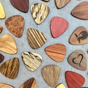 Puede incluir: Una colección de púas de guitarra de madera en varios colores y vetas. Algunas púas tienen patrones únicos, mientras que otras presentan diseños grabados como un corazón o una silueta. Las púas están dispuestas sobre una superficie gris.