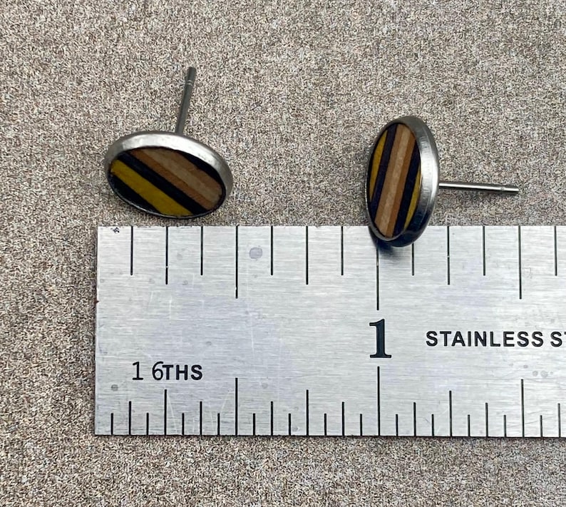 K&ouml;nnte beinhalten: Zwei ovale Ohrstecker mit silberfarbenem Metallrand. Die Ohrringmitten weisen ein gestreiftes Muster aus gelbem, schwarzem und braunem Holz auf. Ein Metalllineal liegt neben den Ohrringen.