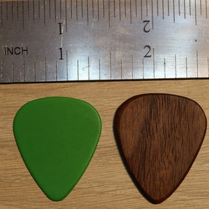 Puede incluir: Dos p&uacute;as de guitarra, una verde y otra marr&oacute;n, se muestran junto a una regla. La p&uacute;a verde es lisa, mientras que la marr&oacute;n tiene un patr&oacute;n de vetas de madera. La regla indica una longitud de aproximadamente 2,5 cm.