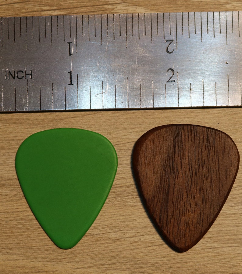 Puede incluir: Dos p&uacute;as de guitarra, una verde y otra marr&oacute;n, se muestran junto a una regla. La p&uacute;a verde es lisa, mientras que la marr&oacute;n tiene un patr&oacute;n de vetas de madera. La regla indica una longitud de aproximadamente 2,5 cm.