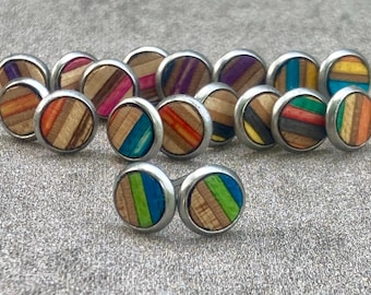 Könnte beinhalten: Eine Sammlung runder Ohrstecker mit silberfarbenem Metallrand. Die Ohrringmitten bestehen aus Holz mit bunten Streifen in verschiedenen Farben, darunter Blau, Grün, Orange und Lila. Die Ohrringe werden auf einer strukturierten grauen Oberfläche präsentiert.