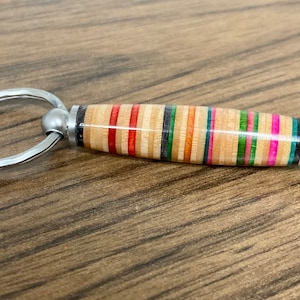 May include: A colorful, striped, wooden keychain with a silver ring. The keychain is made from recycled skateboard wood.