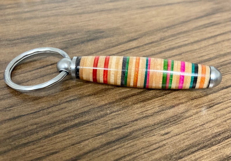 May include: A colorful, striped, wooden keychain with a silver ring. The keychain is made from recycled skateboard wood.