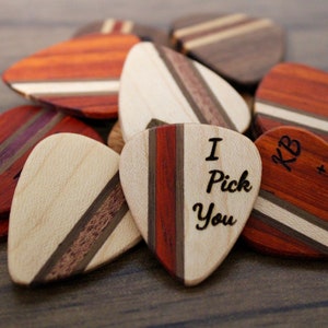 Könnte beinhalten: Ein Stapel aus Holz-Plektronen mit verschiedenen Holzmaserungen und gravierten Botschaften, darunter "I Pick You" und Initialen.