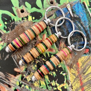 May include: Three colorful keychains made from recycled skateboard wood. Each keychain features a different pattern of wood and is attached to a silver metal key ring.
