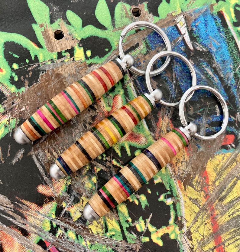 May include: Three colorful keychains made from recycled skateboard wood. Each keychain features a different pattern of wood and is attached to a silver metal key ring.