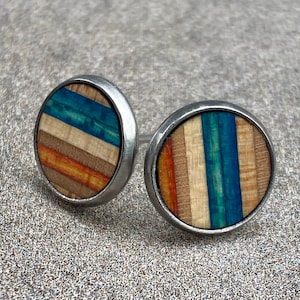K&ouml;nnte beinhalten: Ein Paar runde Ohrstecker mit einem silberfarbenen Metallrahmen. Die Ohrringfl&auml;chen weisen ein farbenfrohes, gestreiftes Design mit Blaut&ouml;nen, Orange und braunem Holz auf. Die Ohrringe sind vor einem strukturierten, grauen Hintergrund platziert.