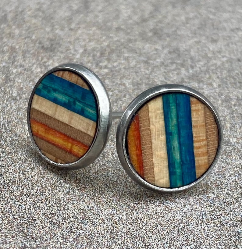 K&ouml;nnte beinhalten: Ein Paar runde Ohrstecker mit einem silberfarbenen Metallrahmen. Die Ohrringfl&auml;chen weisen ein farbenfrohes, gestreiftes Design mit Blaut&ouml;nen, Orange und braunem Holz auf. Die Ohrringe sind vor einem strukturierten, grauen Hintergrund platziert.