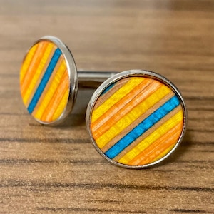 May include: A pair of round cufflinks with a silver border. The cufflinks feature a colorful design of diagonal stripes in yellow, orange, and blue. The cufflinks are made of wood and have a polished finish.