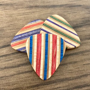 Puede incluir: Tres p&uacute;as de guitarra de madera con dise&ntilde;os de rayas coloridas. Una p&uacute;a presenta rayas verticales rojas, blancas y azules. Las otras dos p&uacute;as tienen rayas horizontales en varios colores, incluyendo azul, verde y rojo. Las p&uacute;as est&aacute;n sobre una superficie de madera.