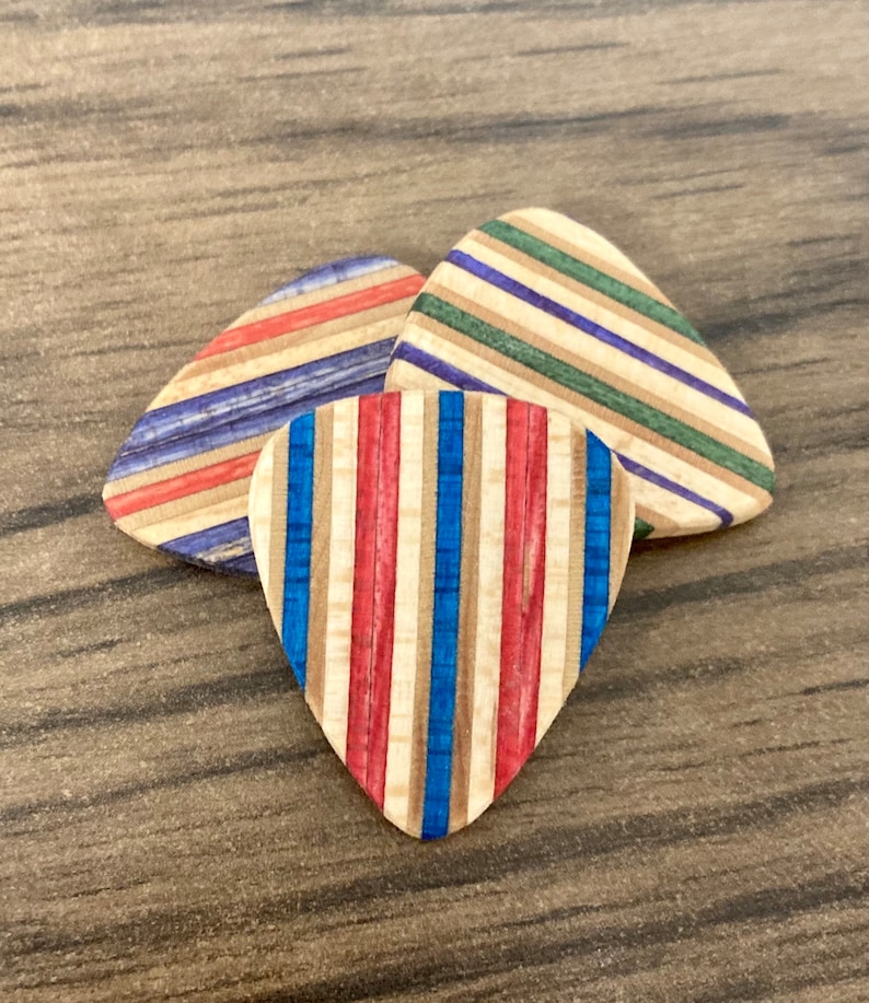 Puede incluir: Tres p&uacute;as de guitarra de madera con dise&ntilde;os de rayas coloridas. Una p&uacute;a presenta rayas verticales rojas, blancas y azules. Las otras dos p&uacute;as tienen rayas horizontales en varios colores, incluyendo azul, verde y rojo. Las p&uacute;as est&aacute;n sobre una superficie de madera.
