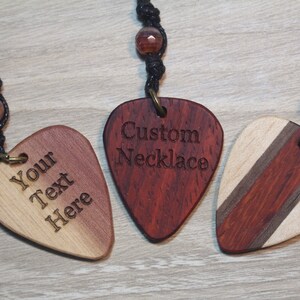 Puede incluir: Tres collares de p&uacute;as de guitarra de madera con cordones negros y cuentas decorativas. Uno tiene el texto "Your Text Here", otro dice "Custom Necklace", y el tercero tiene un dise&ntilde;o a rayas.