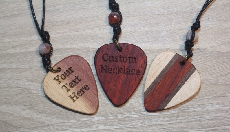 Puede incluir: Tres collares de p&uacute;as de guitarra de madera con cordones negros y cuentas decorativas. Uno tiene el texto "Your Text Here", otro dice "Custom Necklace", y el tercero tiene un dise&ntilde;o a rayas.