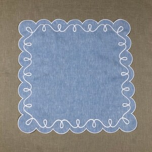 May include: A blue linen table runner with a white scalloped edge. The edge is decorated with a continuous white embroidered design.