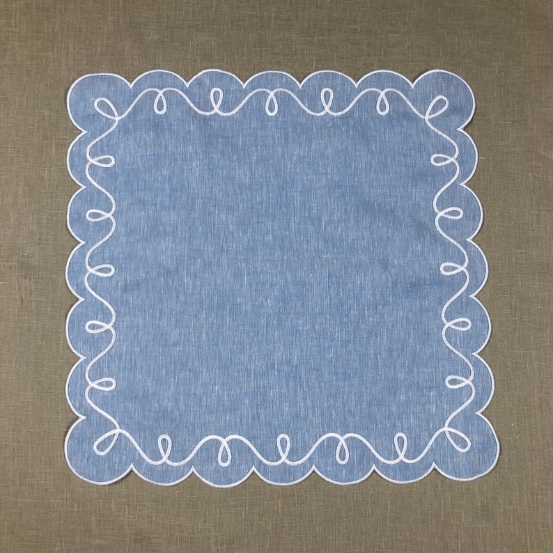 May include: A blue linen table runner with a white scalloped edge. The edge is decorated with a continuous white embroidered design.