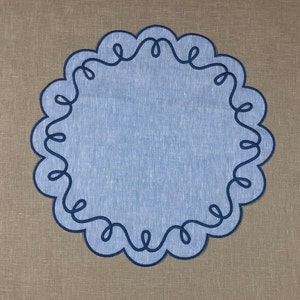 May include: A blue linen placemat with a decorative blue embroidered border. The border is a continuous design of loops and swirls.