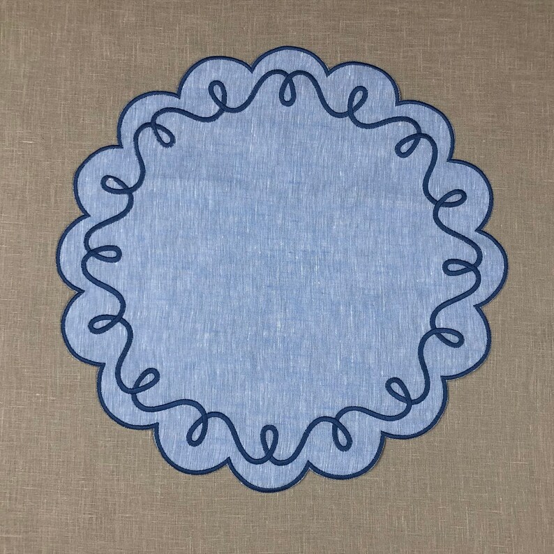 May include: A blue linen placemat with a decorative blue embroidered border. The border is a continuous design of loops and swirls.