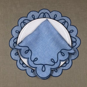 May include: Blue linen napkin with a scalloped edge and a decorative design. The napkin is placed on a white plate with a blue and white scalloped edge.